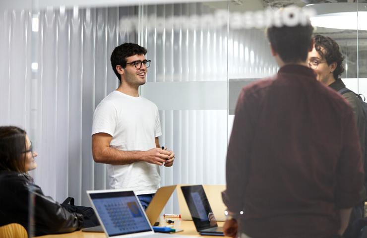 A young man in a white t-shirt is engaged in conversation with people in a modern office environment.