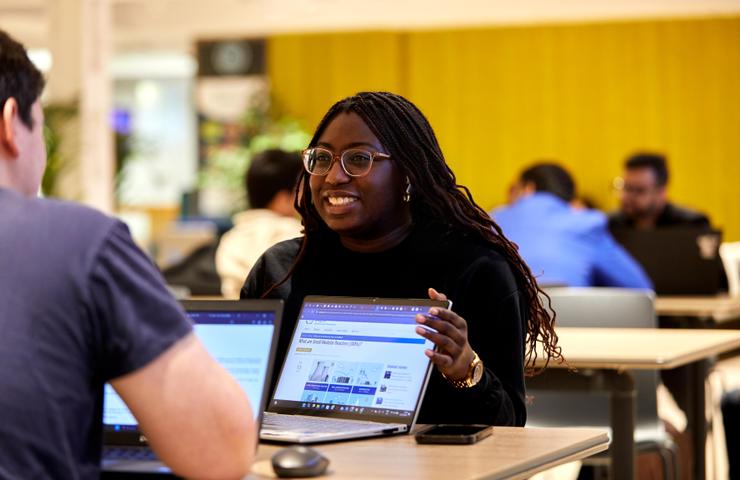 A woman with glasses discussing over a laptop in a busy office area.