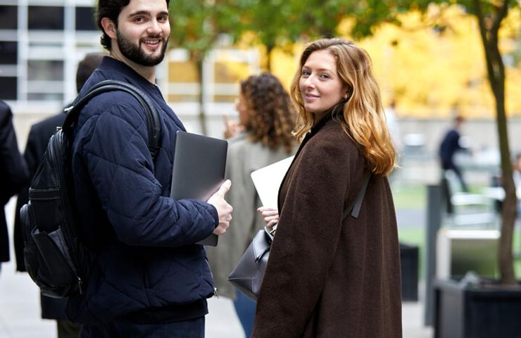 Two young professionals smiling and looking back while walking in a city setting with laptops in hand.