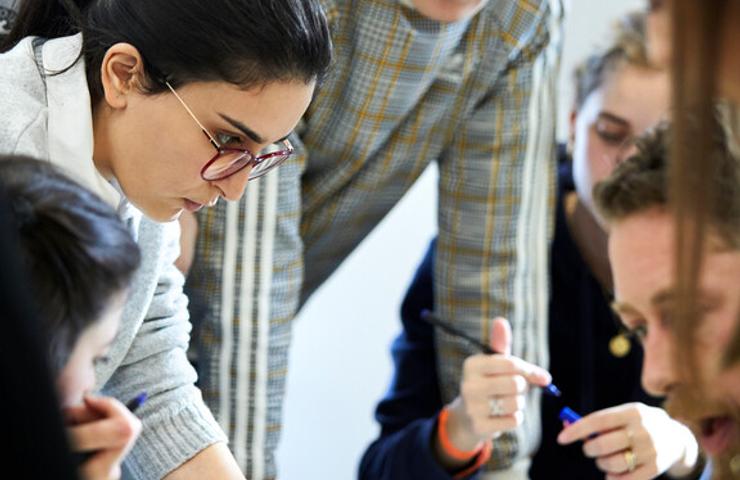 A group of young adults are engaged in discussion and examining documents around a table.