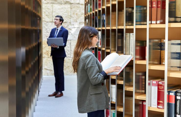 A woman and a man, dressed in business attire, are reading books in a library with floor-to-ceiling shelves filled with books.