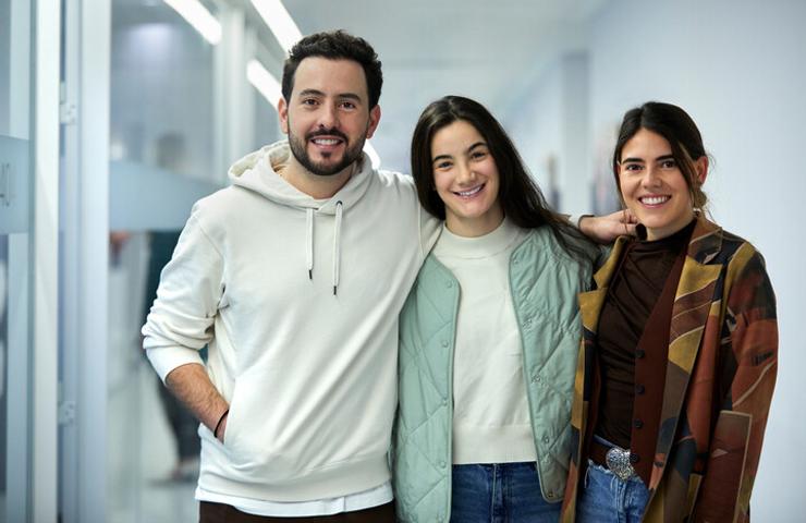 Three young adults, two women and one man, smiling and posing together indoors.