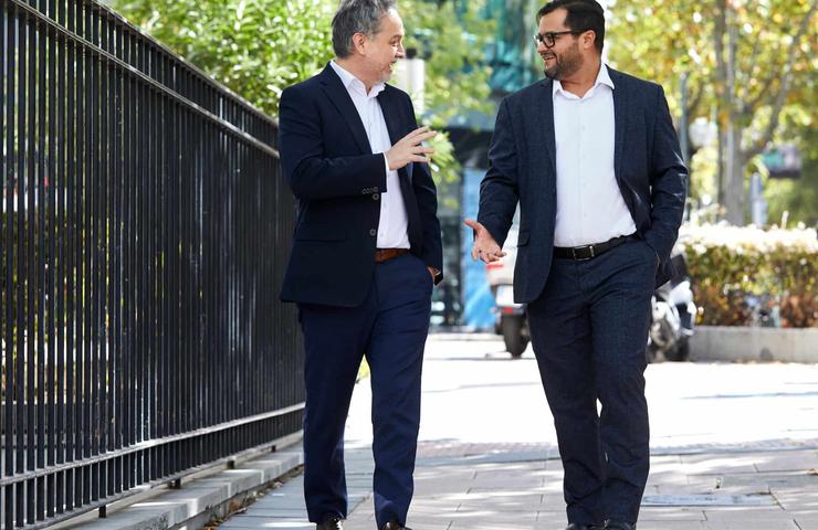 Two men in business attire are walking and chatting happily on a sunny street.
