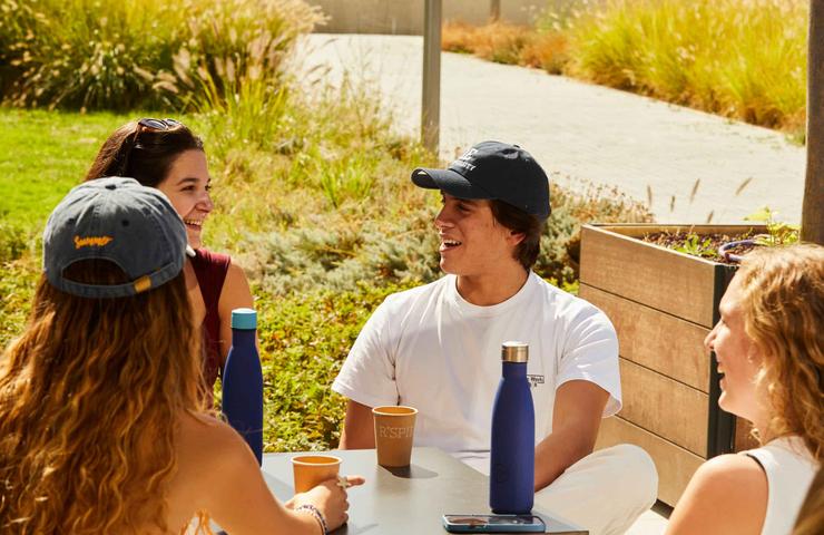 Four young adults enjoying a conversation at an outdoor table with coffee and water bottles.