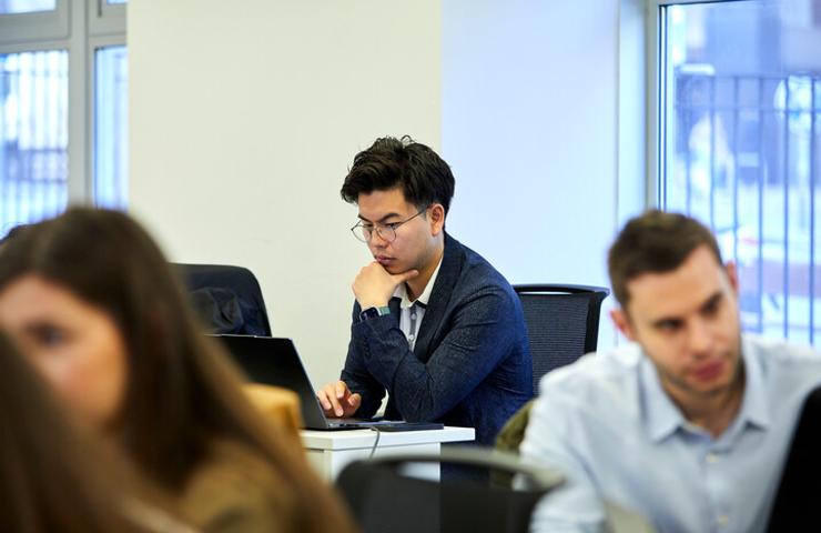 A young man focused on his laptop in a busy classroom setting.