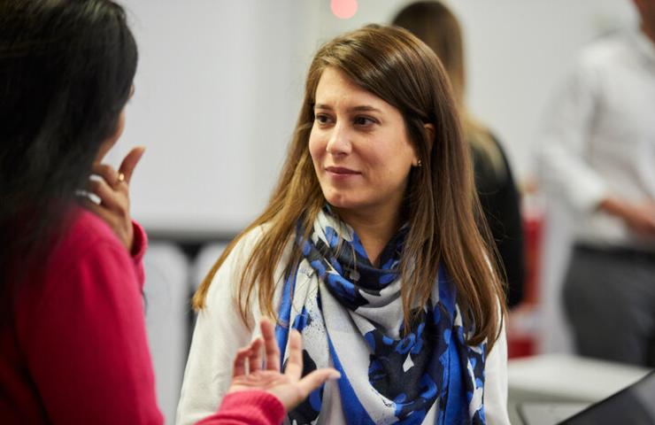 Two women engaged in a conversation in a busy office setting.