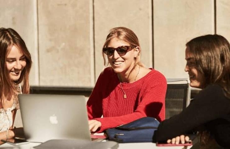 Three women sit at a table, smiling and talking with a laptop open in front of them.