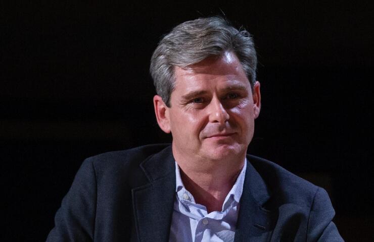 A middle-aged man with gray hair, wearing a blazer and a checkered shirt, looks pensive and slightly frowning in a conference setting.