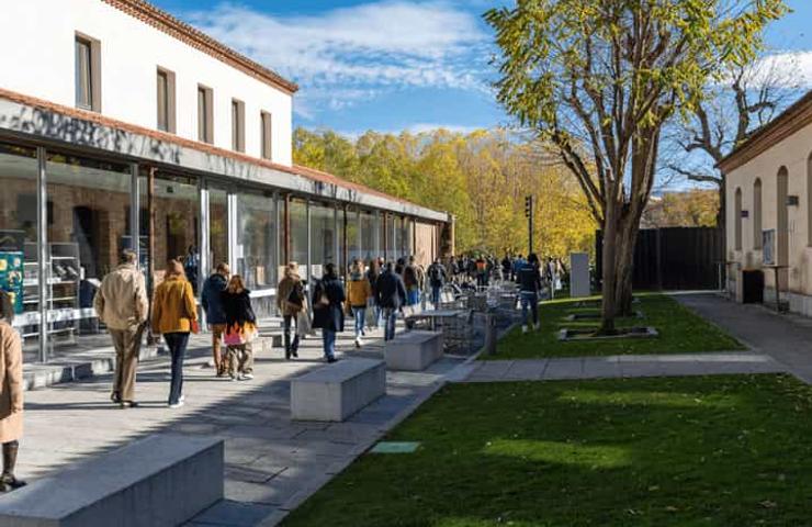 People walking near modern glass building in a park setting with trees and blue sky.