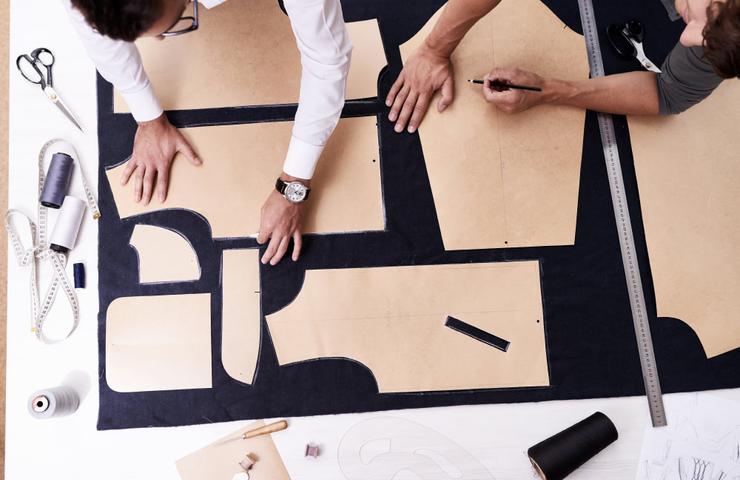 Two people working on tailoring patterns on a large piece of fabric, with measuring tools and scissors around them.