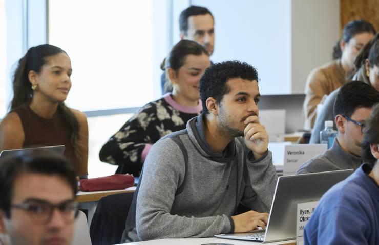 A diverse group of students attentively listening in a classroom setting, with laptops open and some taking notes.