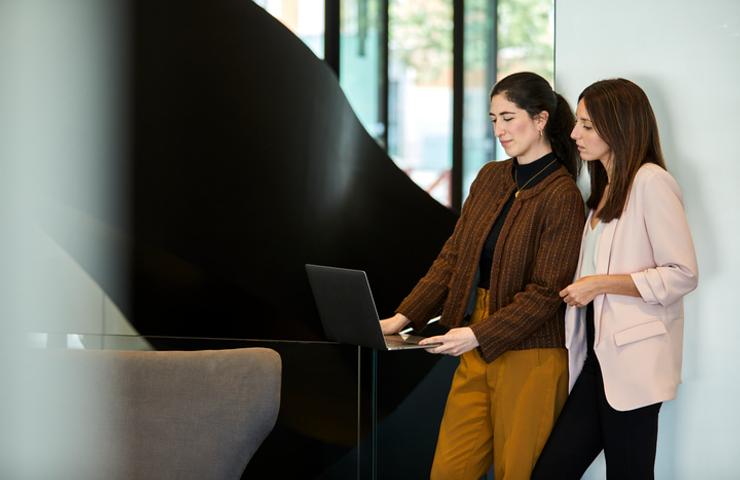 Two women standing by a laptop in a modern office environment, engaging in a discussion.