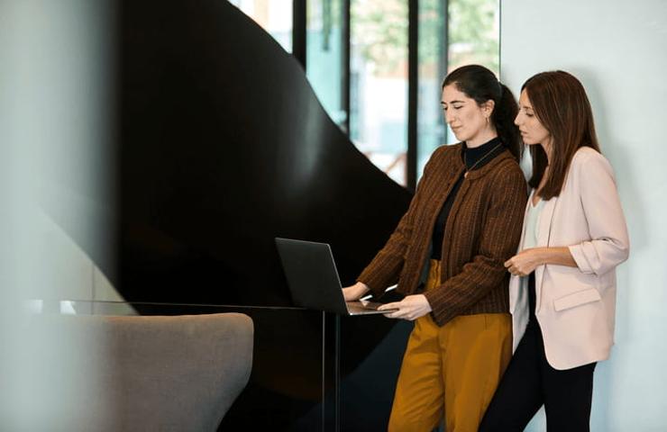 Two women standing and looking at a laptop in a modern office setting.