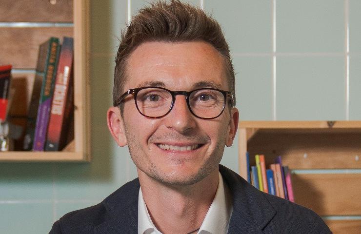 A man with glasses smiling in a room with a bookshelf in the background.