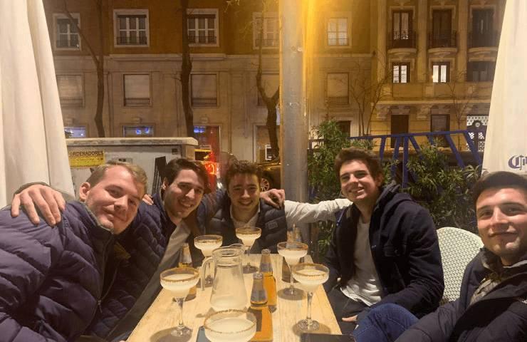 A group of five young men smiling at a table with drinks in an outdoor dining area at night.