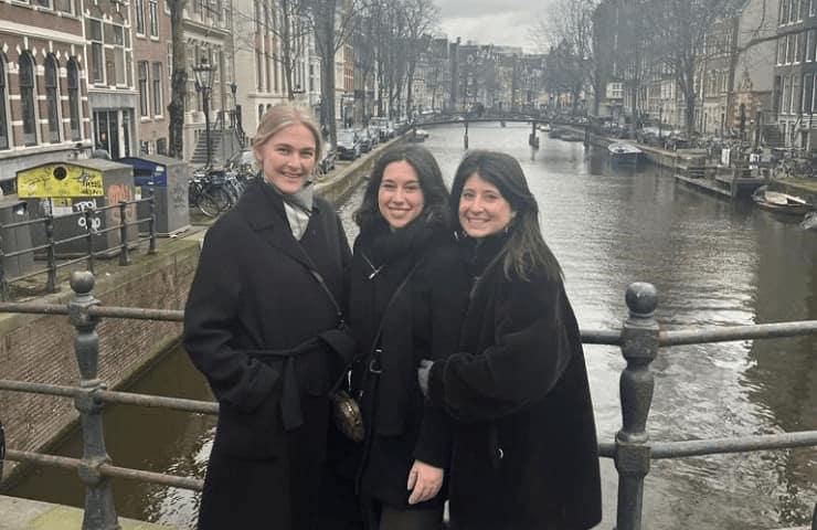 Three women smiling in front of a canal in an urban setting.