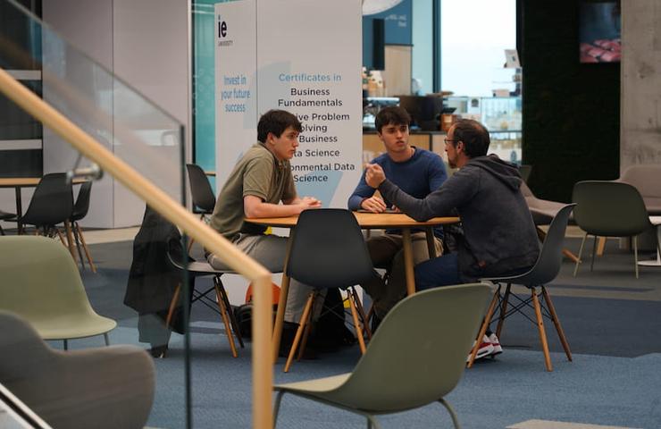 Three people are sitting at a table, having a discussion in a modern indoor setting with various chairs and banners related to business and data science.