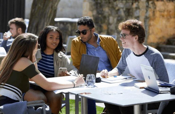 A group of young adults engaging in a discussion at an outdoor café table, with laptops and smartphones visible.