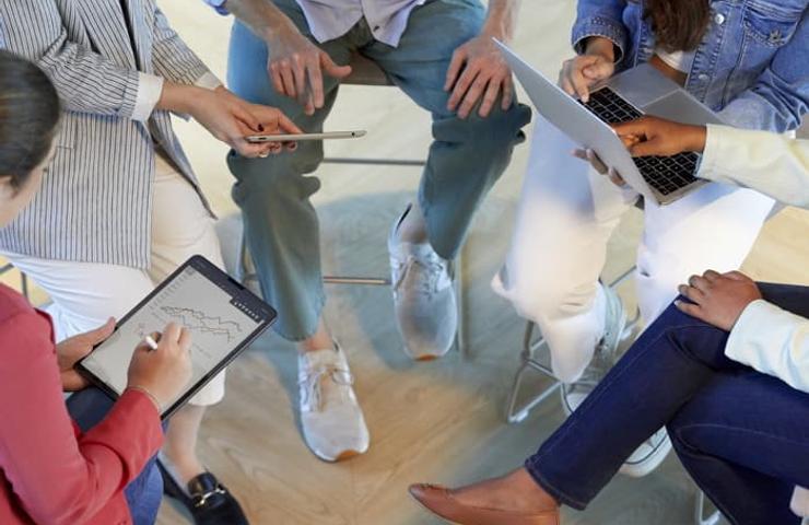 A group of people sitting in a circle, engaged in a discussion with laptops and digital tablets.