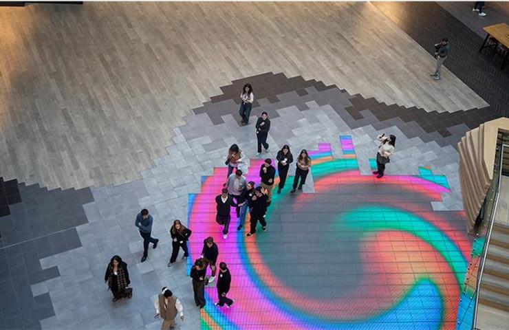 A colorful digital art installation on the floor with people gathered around it.