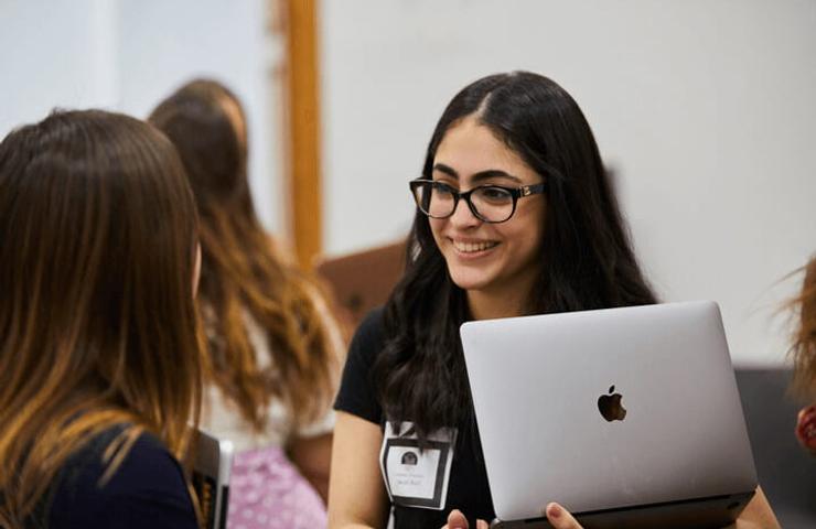 A young woman with glasses is smiling and talking to her peers while holding a laptop at a gathering.