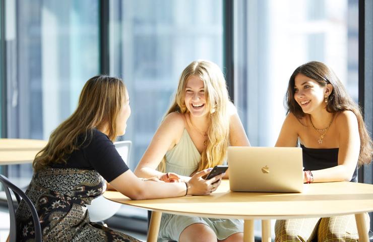 Three young women are sitting together at a table, engaging in a lively conversation with a laptop in front of them.