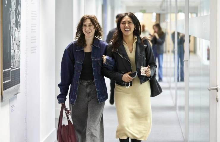 Two women walking and laughing together in a gallery with photographs on the walls.