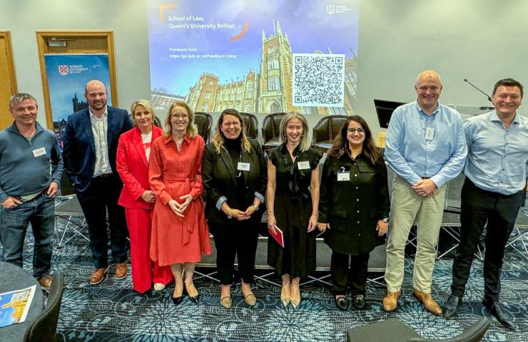 A group of nine individuals posing for a photo at an event, with a presentation screen displaying a QR code and the name 'School of Law, Queen's University Belfast' in the background.