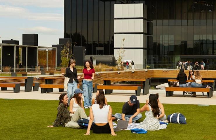 A group of young people sitting and standing on a grassy area at a modern university campus.