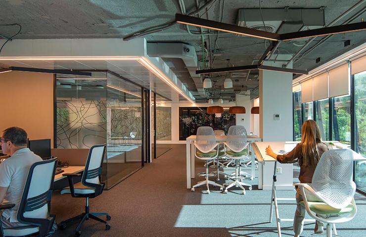 An office space with individuals working at desks equipped with computers, in a modern setup with glass walls and exposed ceiling.