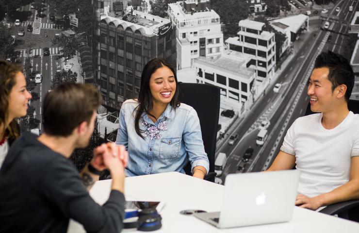 Four people, two men and two women, are engaged in a cheerful conversation around a table with a laptop in an office setting with a cityscape mural in the background.