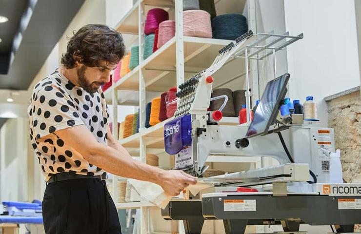 A man in a polka dot shirt operates a modern embroidery machine in a colorful workshop.