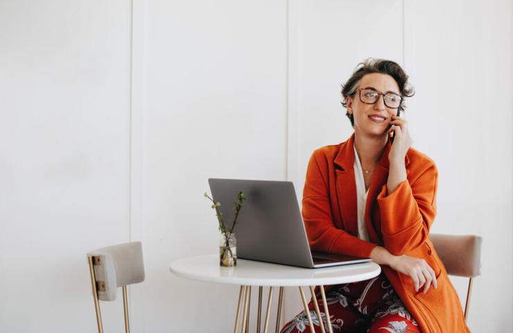 A smiling woman with glasses, sitting by a laptop at a white table, dressed in a bright orange cardigan and a floral skirt, talking on the phone.