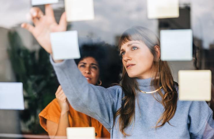 Two women collaborating on ideas using sticky notes on a glass window in an office setting.