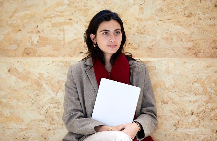 A young woman in a beige coat and red scarf sits thoughtfully while holding a blank notepad.