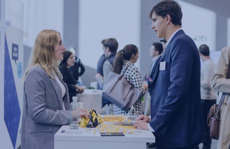 A man and a woman having a conversation at a promotional booth in a busy conference hall.