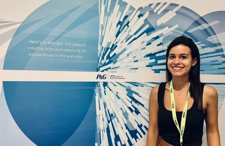 A woman smiling in front of a corporate anniversary banner that reads 'Here's to the next 175 years of creating firsts and unlocking the extraordinary in the everyday.'