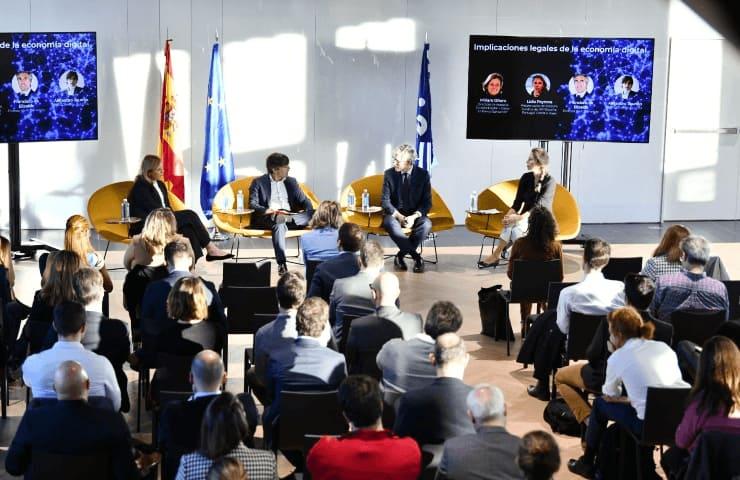 A conference setting with five speakers seated on stage in discussion in front of an audience.