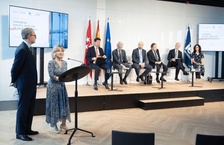 A woman is speaking at a podium in a conference room with several attendees seated behind her under national flags.