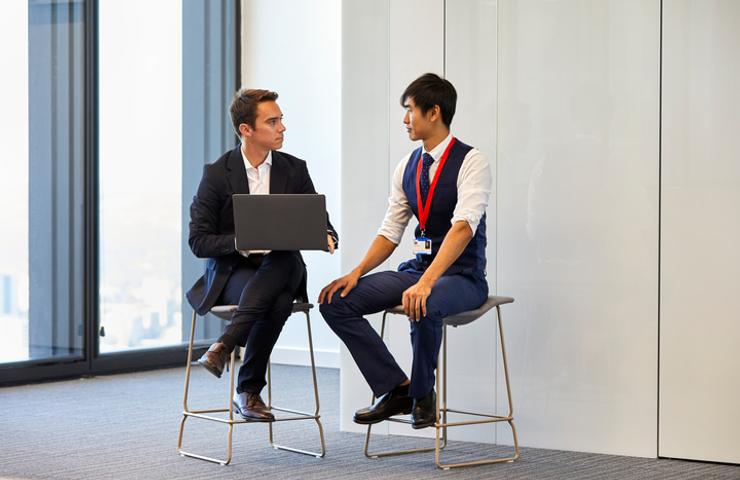 Two men in business attire are having a discussion while sitting on stools in an office with a city view.