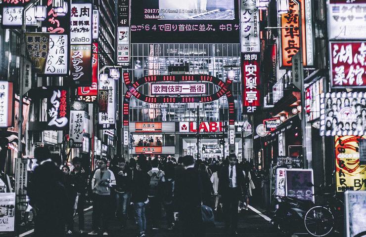 A bustling street scene at night in a Japanese city, illuminated by numerous neon signs and filled with people.