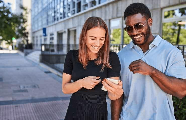 A man and a woman are smiling and looking at a smartphone together on a sunny city street.