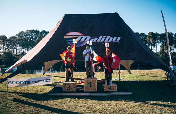 Three athletes stand on a podium at an outdoor sporting event under a large tent with the Malibu logo.