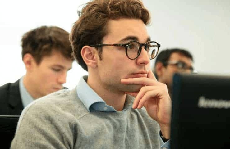 A young man with glasses is focused on a computer screen in a classroom setting.