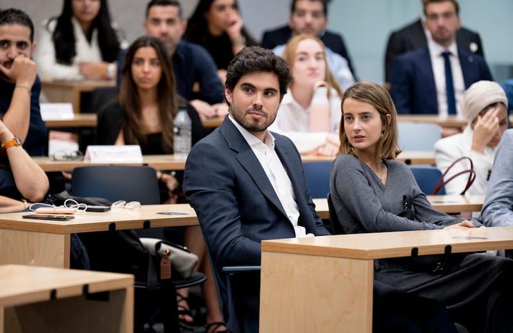 A young man and woman sitting together in a lecture hall filled with students, appearing focused and attentive.