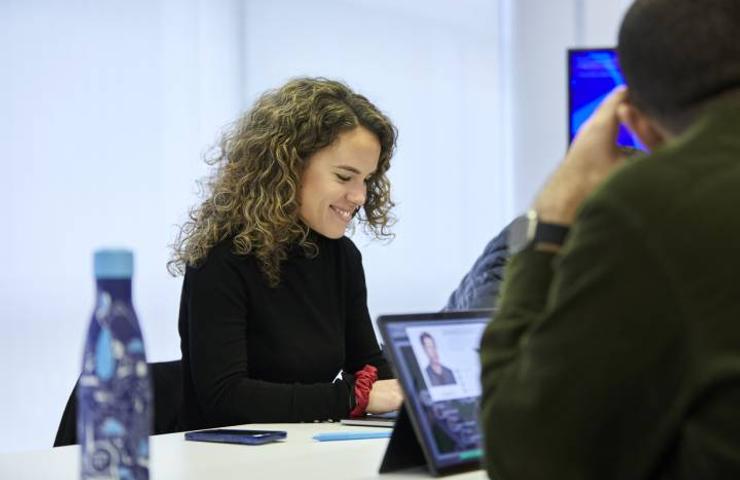 A woman with curly hair is smiling while using a laptop at a table in a bright environment.