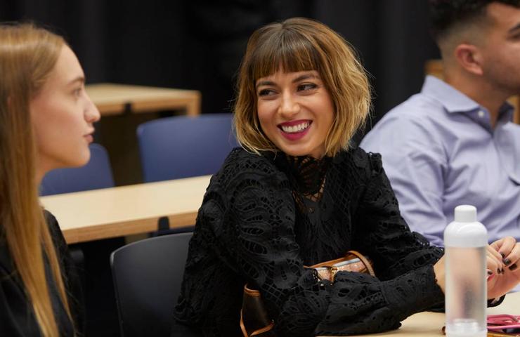 A woman with a bob haircut and black lace top smiles engagingly while sitting at a table with two other people in a conference or classroom setting.