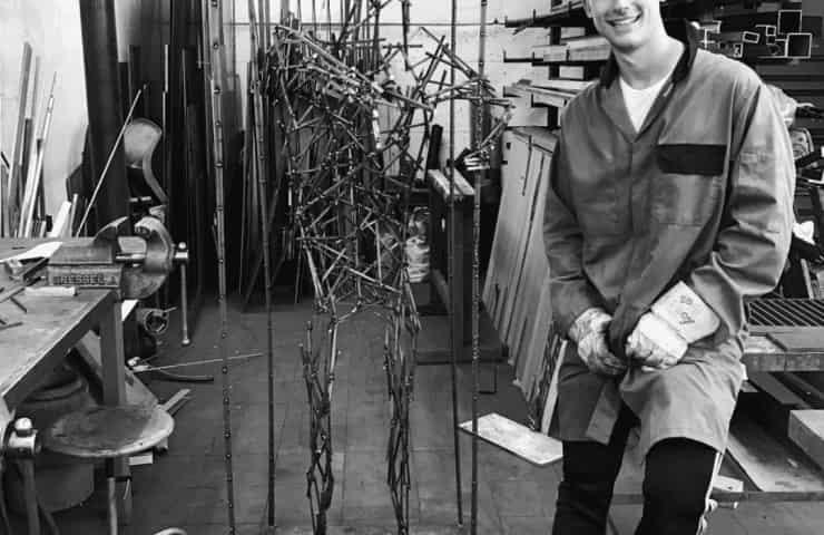 A young man in a workshop wearing a welding jacket, smiling proudly next to a metal sculpture.
