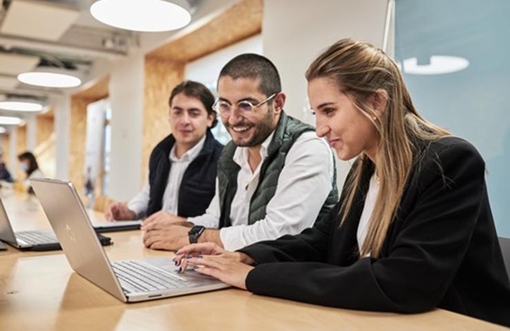 A group of three young professionals collaborating on a laptop in a modern office environment.