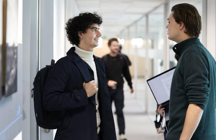 Two men talking in a corridor with one holding a backpack and the other holding documents.